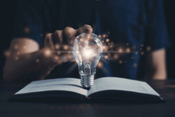 A person touching a glowing light bulb emerging from an open book, symbolizing inspiration and creativity.