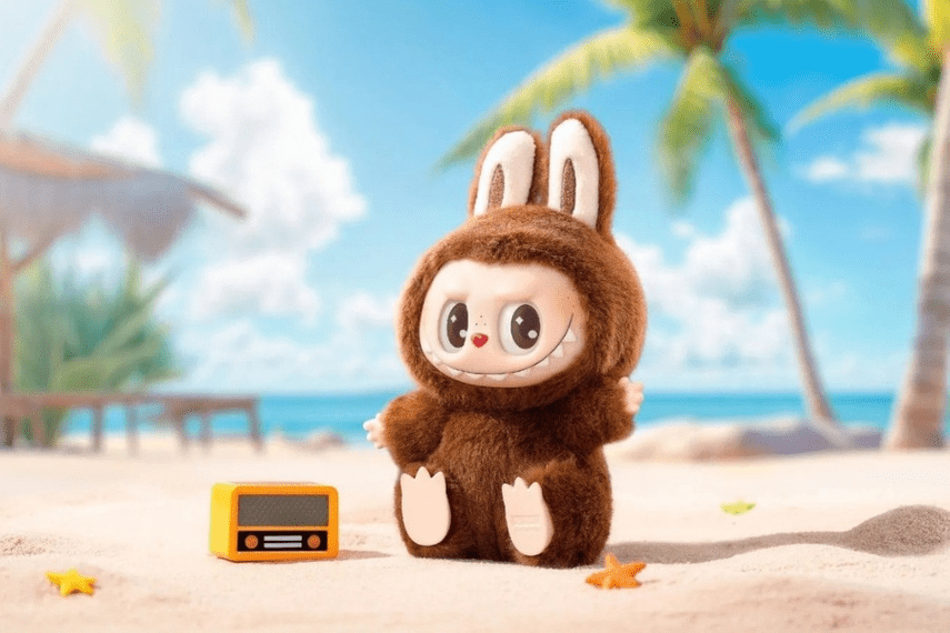 A cute, furry Labubu doll dressed in a brown rabbit costume, featuring big ears and a playful smile, sitting on a sandy beach with blue water and palm trees in the background.