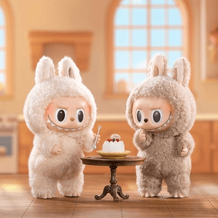 Two cute, plush monster dolls wearing bunny suits, standing next to a small cake on a table in a cozy kitchen setting.