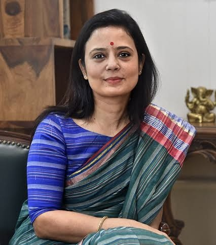 A woman wearing a traditional saree, smiling at the camera while seated in an indoor setting with a decorative background.