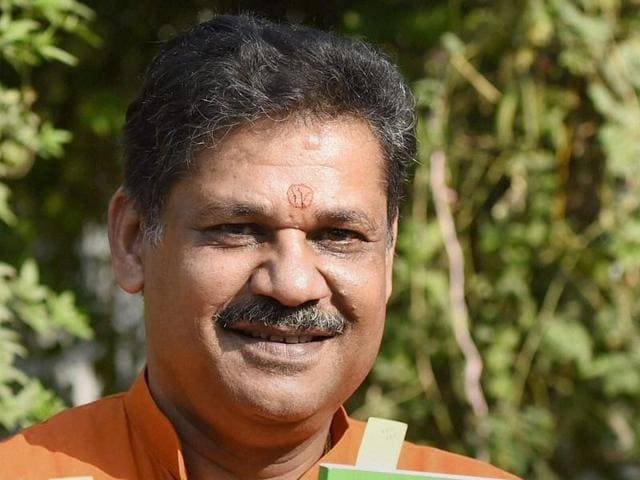A man smiling at the camera, wearing an orange shirt, with a tilak mark on his forehead, surrounded by greenery.